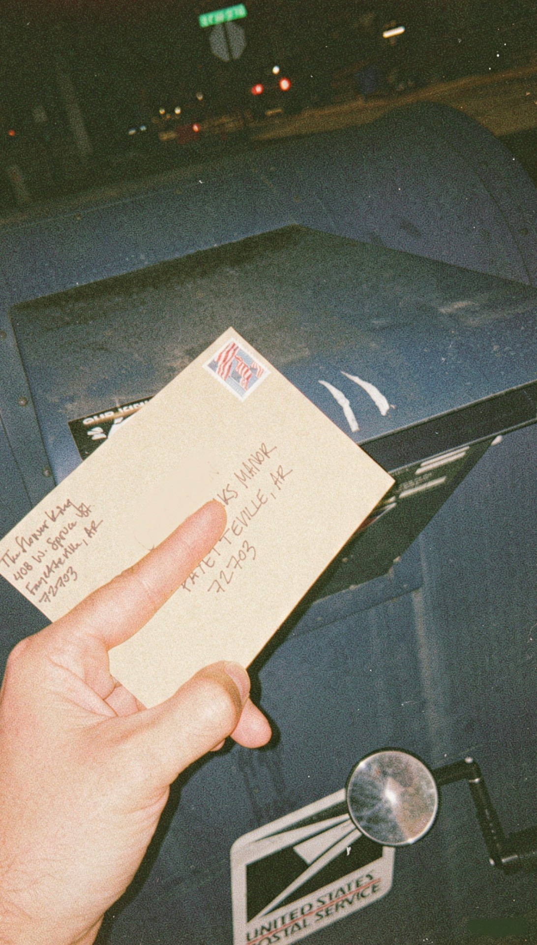 A hand holding a stamped, hand-addressed kraft envelope at a blue USPS mailbox at night