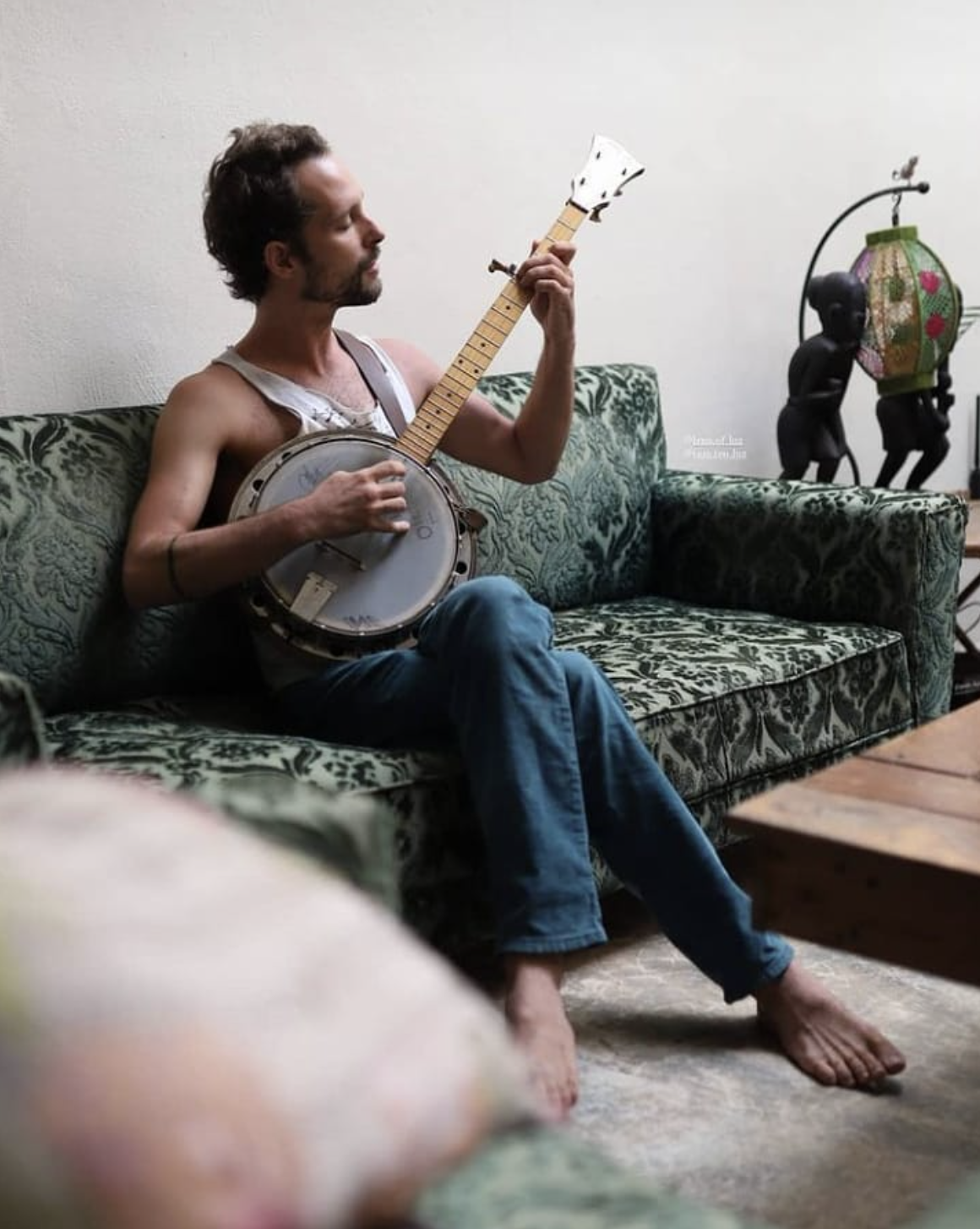 Teo Luz sitting barefoot on a green velvet couch, playing a banjo